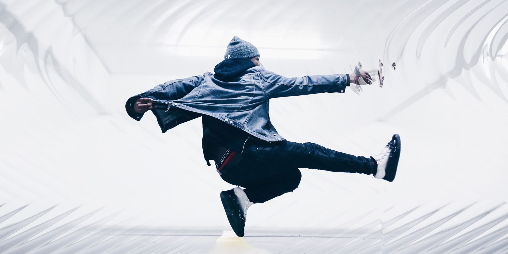 Energetic dancer mid-air in a modern dance pose, wearing a denim jacket and beanie, set against a minimalist white background.